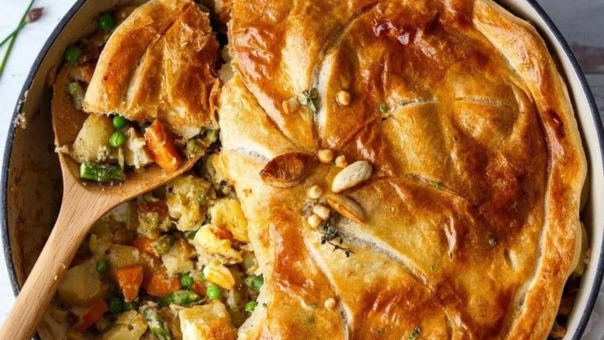 A vegetable pot pie with a golden-brown crust, partially cut open to reveal a filling of carrots, peas, potatoes, and other vegetables. A wooden spoon scoops out a portion.