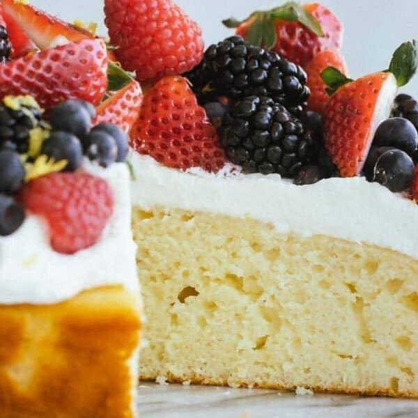 A slice of vanilla cake with white frosting, topped with fresh strawberries, blackberries, blueberries, and raspberries.