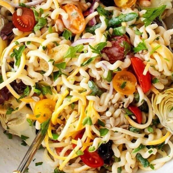 A bowl of pasta salad containing cherry tomatoes, artichoke hearts, olives, and fresh herbs, with two serving utensils.