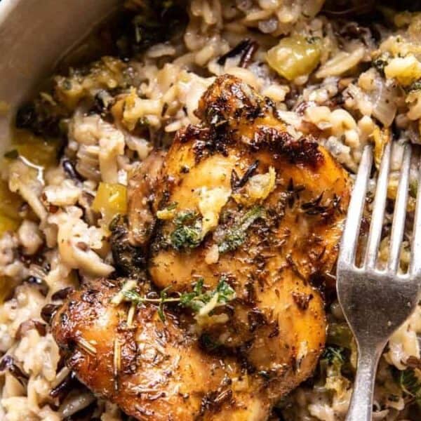 A bowl of wild rice with a grilled chicken thigh on top, garnished with herbs, accompanied by a fork and spoon.