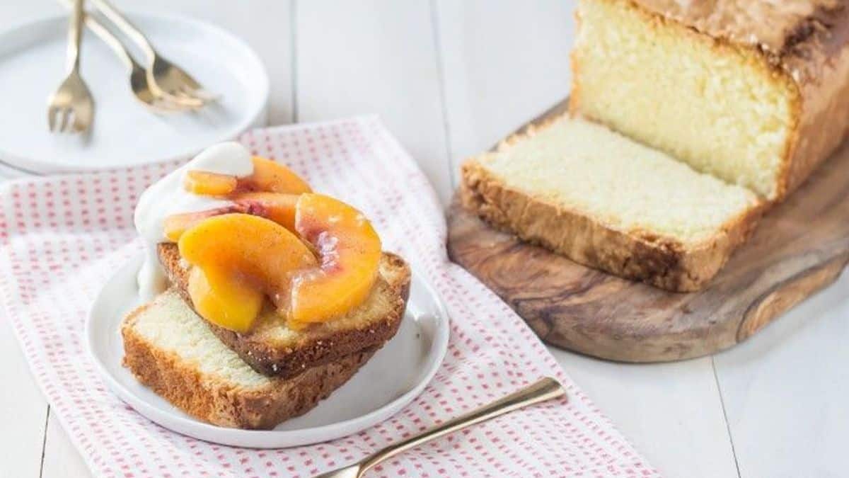 A white plate with a slice of pound cake topped with peach slices and whipped cream. Next to the plate is a loaf of pound cake resting on a wooden board.