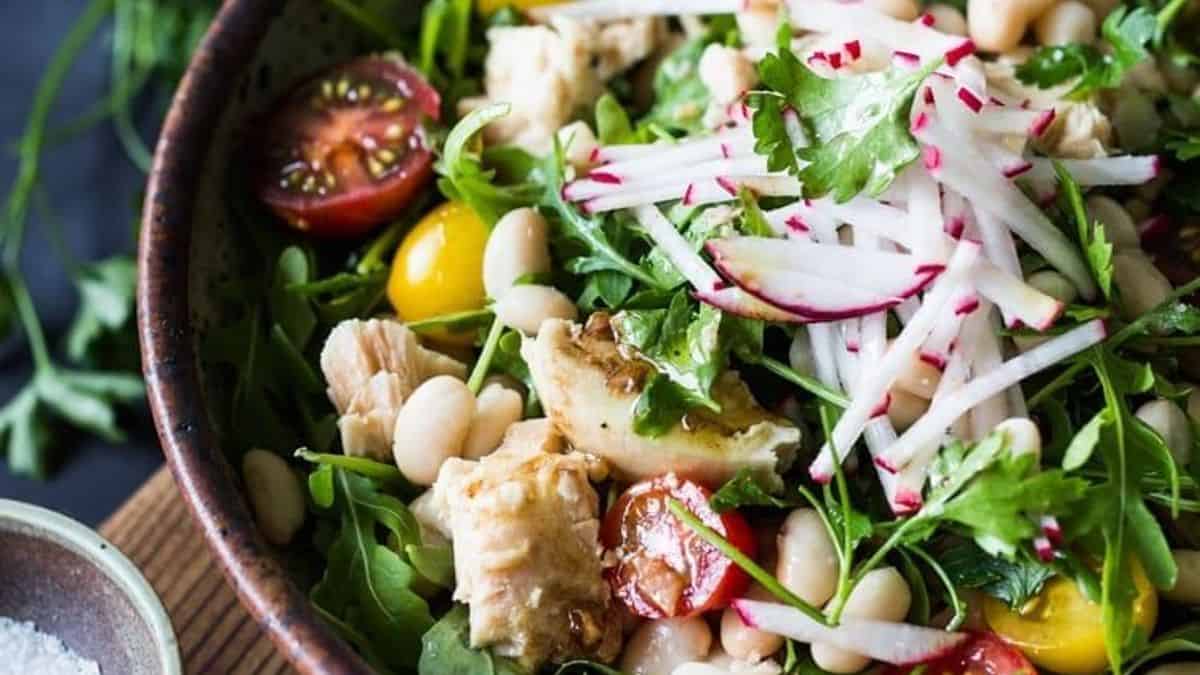 A bowl containing mixed greens, cherry tomatoes, white beans, grilled chicken slices, sliced radishes, and cilantro.
