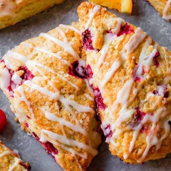 Triangular scones with a white icing drizzle, garnished with orange slices and whole cranberries, displayed on a gray surface.