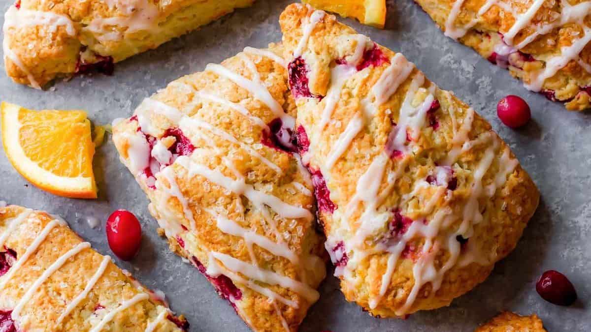 Triangular scones with a white icing drizzle, garnished with orange slices and whole cranberries, displayed on a gray surface.
