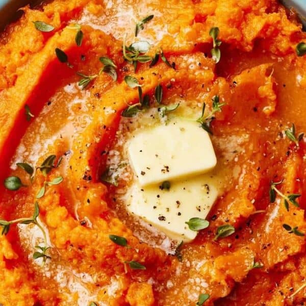 A bowl of mashed sweet potatoes topped with two pats of butter, black pepper, and fresh herbs.