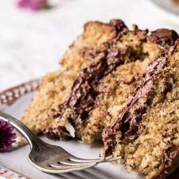 A slice of layered cake with chocolate frosting sits on a ceramic plate. A fork rests beside the cake, and a small purple flower is placed near it.