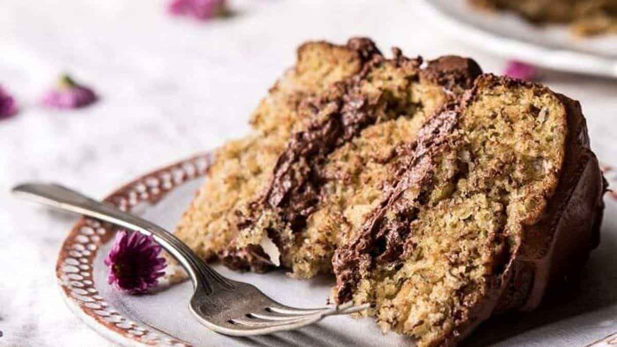 A slice of layered cake with chocolate frosting sits on a ceramic plate. A fork rests beside the cake, and a small purple flower is placed near it.