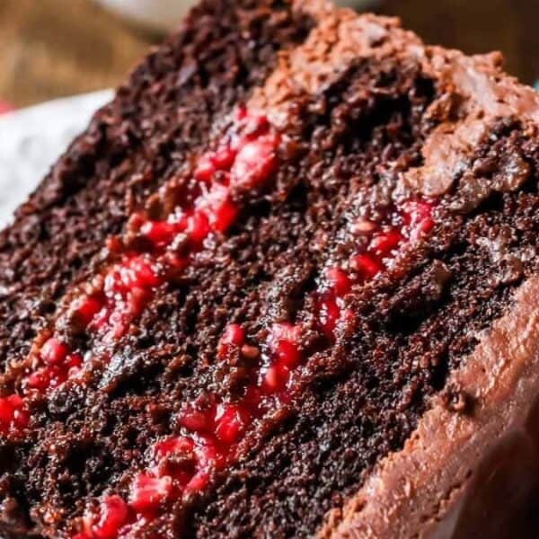 A slice of chocolate cake with chocolate frosting and raspberry filling sits on a white plate, topped with fresh raspberries.