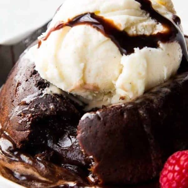 A chocolate lava cake topped with a scoop of vanilla ice cream and drizzled with chocolate sauce, accompanied by fresh raspberries on a white plate.