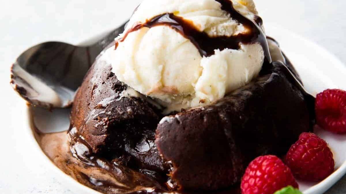 A chocolate lava cake topped with a scoop of vanilla ice cream and drizzled with chocolate sauce, accompanied by fresh raspberries on a white plate.