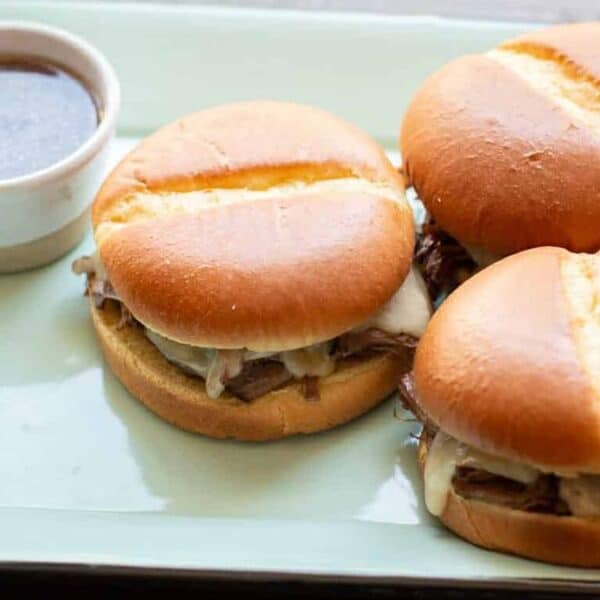 Three roast beef sandwiches with melted cheese on buns are placed on a tray beside a small cup of au jus dipping sauce.