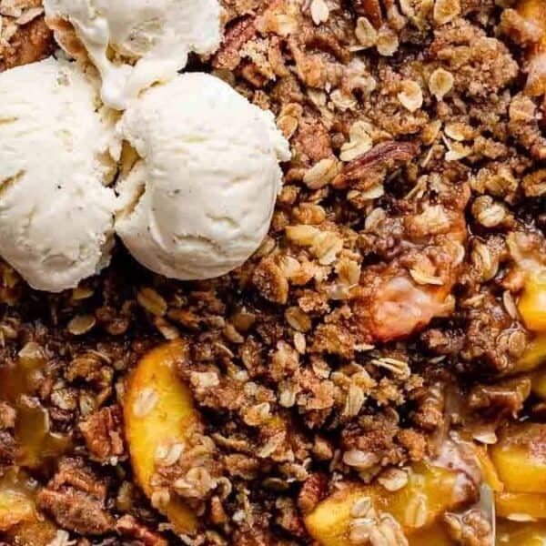 A white dish containing a baked peach dessert with an oat crumble topping and two scoops of vanilla ice cream.