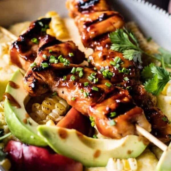 A bowl containing grilled chicken skewers, sliced avocado, corn, fresh cilantro, chives, and creamy yellow polenta.