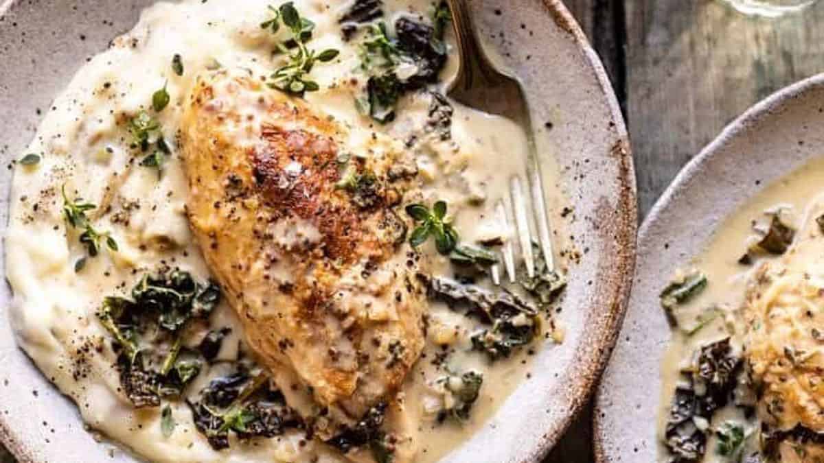 A plate with sliced cooked chicken breast, creamy sauce, wilted greens, and a sprinkle of fresh herbs on top.