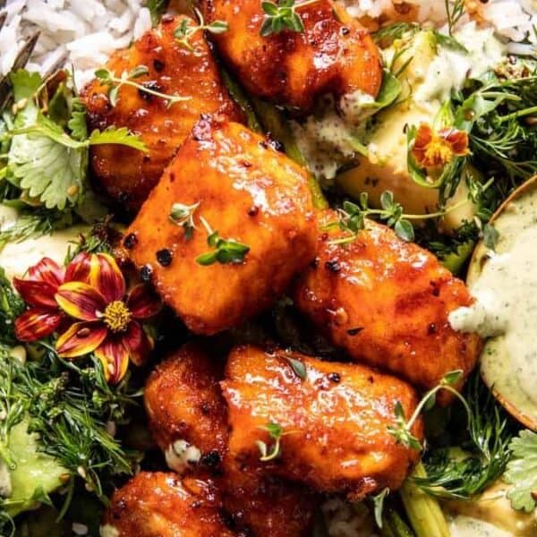 A bowl of rice topped with glazed chicken pieces, fresh green herbs, edible flowers, a portion of green sauce on the side, and a fork placed next to the bowl.