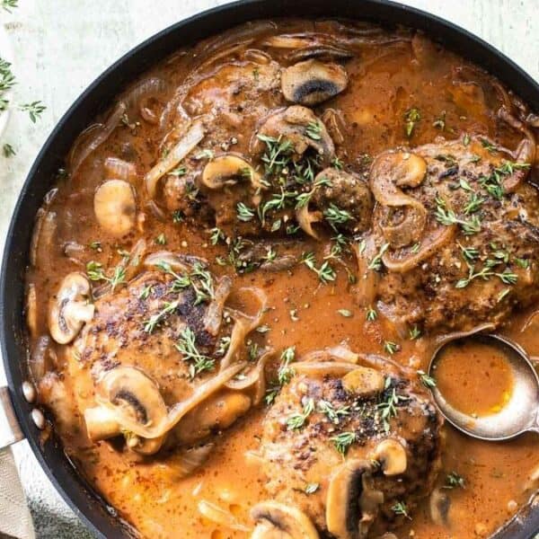 A skillet containing chicken thighs in a brown mushroom and onion sauce, garnished with fresh thyme. A spoon is in the sauce and a small bowl of thyme sits nearby.