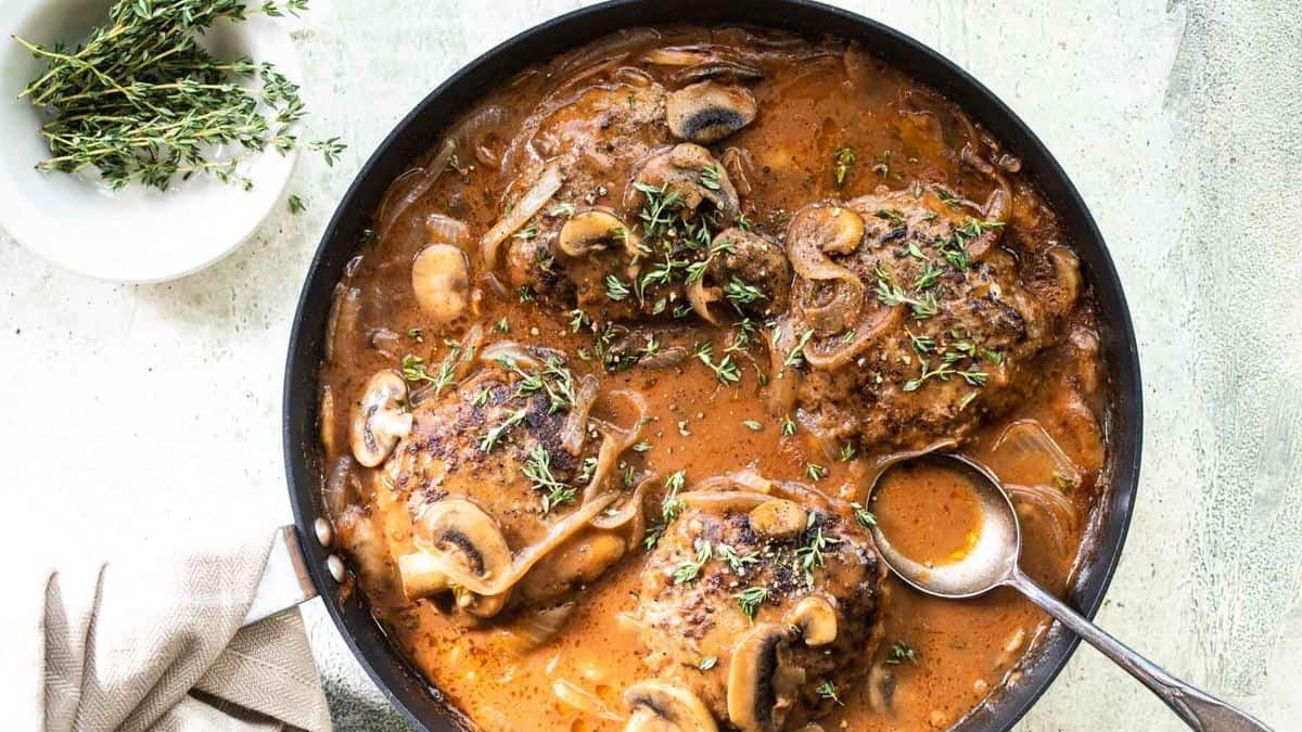 A skillet containing chicken thighs in a brown mushroom and onion sauce, garnished with fresh thyme. A spoon is in the sauce and a small bowl of thyme sits nearby.