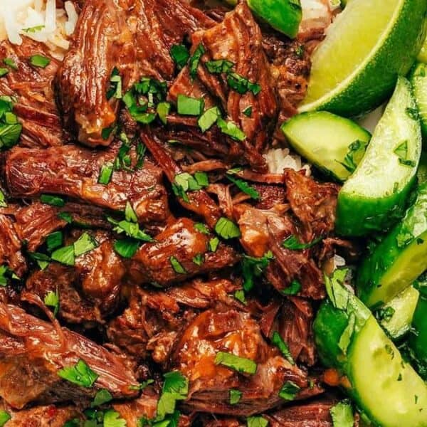 A bowl containing white rice, shredded beef, sliced cucumbers, lime wedges, and chopped herbs.