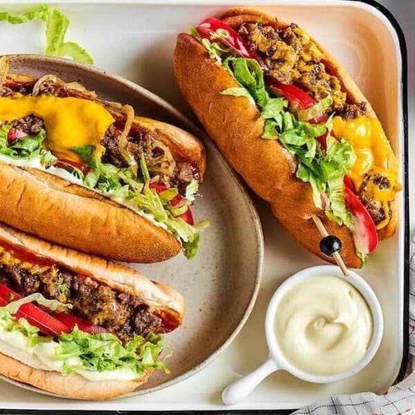 Three hot dogs topped with ground beef, shredded cheese, chopped lettuce, diced tomato, and onions are arranged on a white tray alongside a serving of mayonnaise and shredded lettuce.