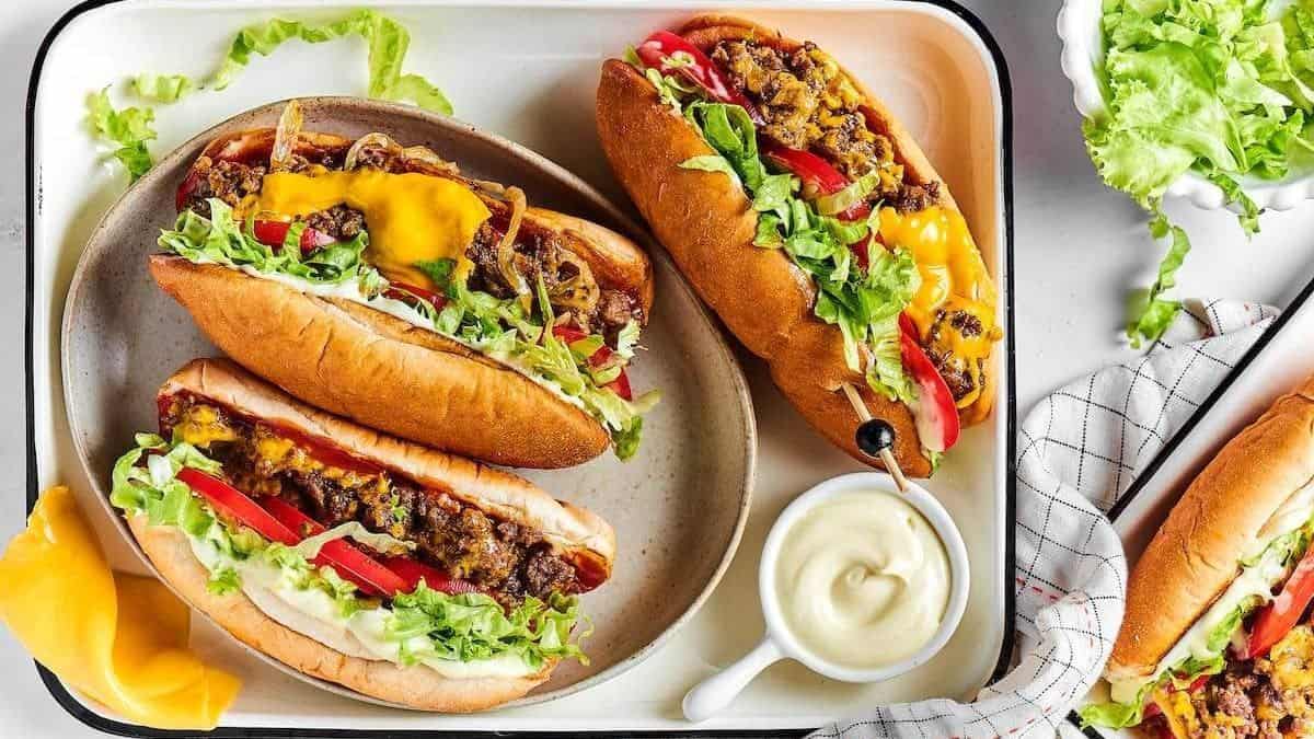 Three hot dogs topped with ground beef, shredded cheese, chopped lettuce, diced tomato, and onions are arranged on a white tray alongside a serving of mayonnaise and shredded lettuce.