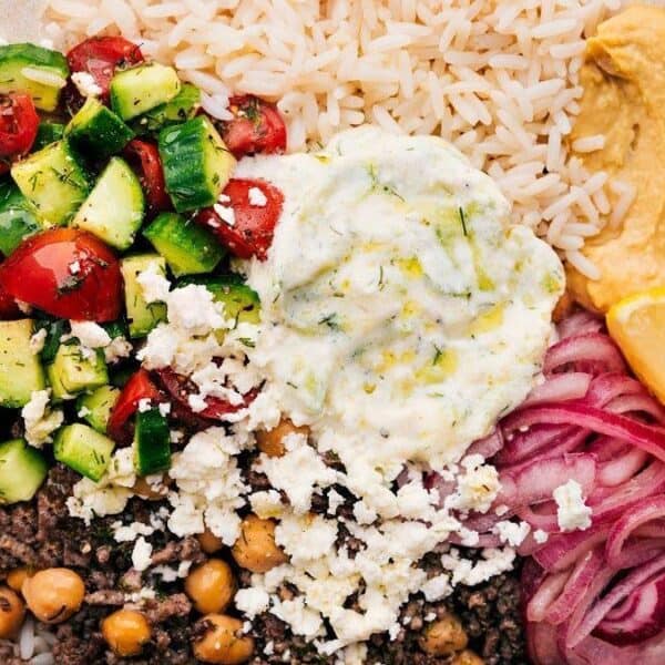 A bowl containing rice, hummus, parsley, a lemon wedge, pickled onions, ground beef with chickpeas, diced cucumber and tomato salad, tzatziki sauce, and crumbled feta cheese.