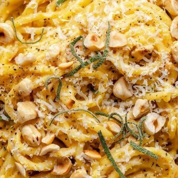 A plate of pasta topped with grated cheese, chopped hazelnuts, fresh herbs, and black pepper, with a spoon beside it.