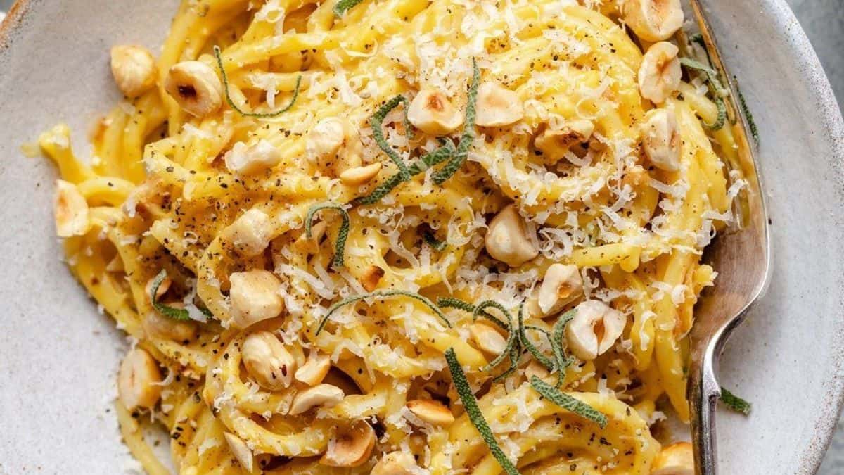 A plate of pasta topped with grated cheese, chopped hazelnuts, fresh herbs, and black pepper, with a spoon beside it.