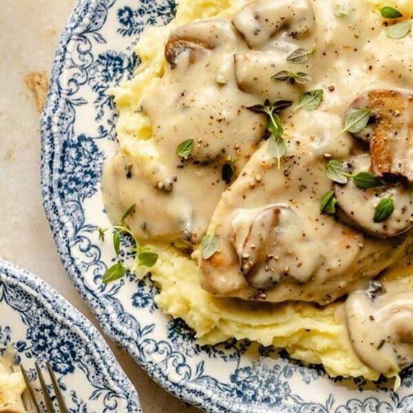 Mashed potatoes topped with sliced chicken breast, creamy mushroom sauce, and fresh herbs on a blue and white patterned plate.
