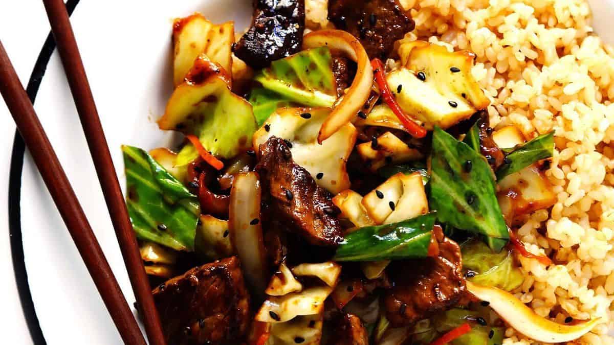 A plate with stir-fried beef, cabbage, onions, and bell peppers alongside a serving of brown rice; chopsticks are placed beside the plate.