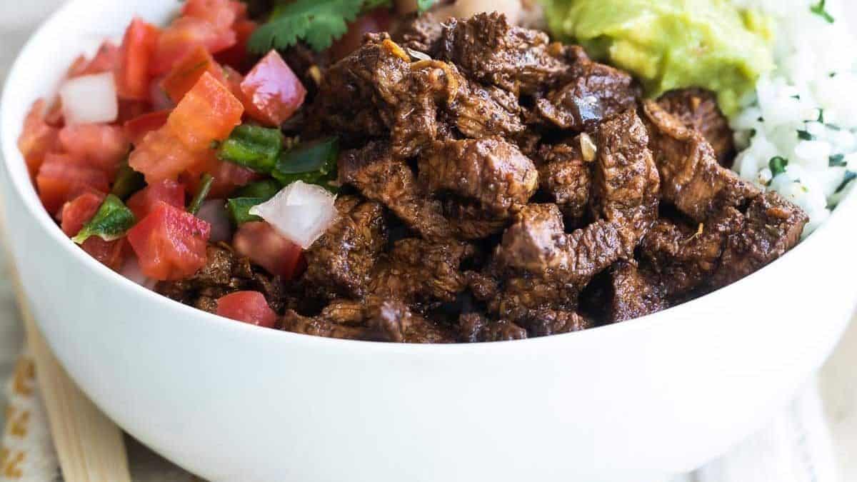 A white bowl containing seasoned steak, diced tomatoes, cilantro-lime rice, pinto beans, and guacamole.