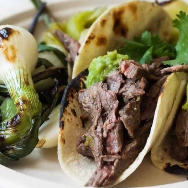 Two steak tacos topped with guacamole and cilantro, served with grilled green onions on a white plate.