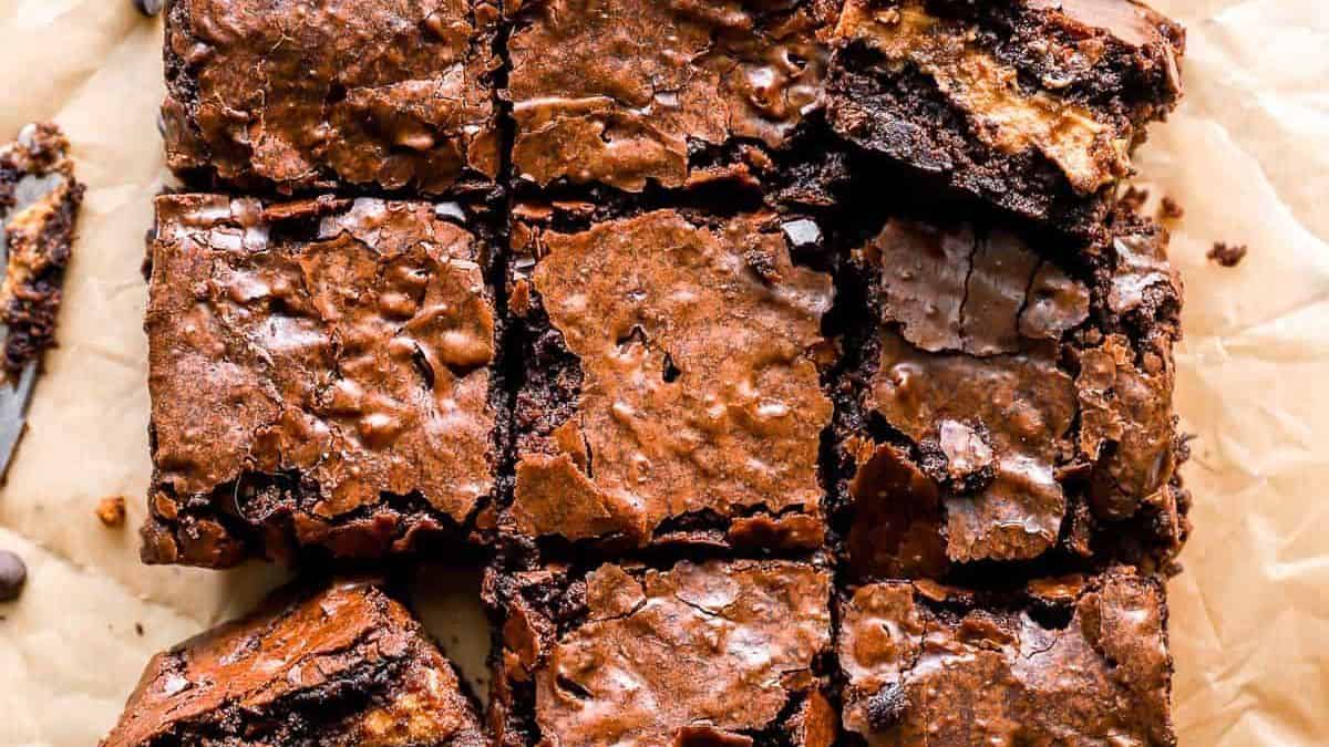 Nine square chocolate brownies with cracked, shiny tops are arranged on parchment paper.