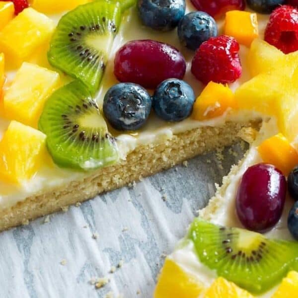 A round dessert with a cream-covered crust, topped with sliced strawberries, blueberries, kiwi slices, grapes, raspberries, and pieces of mango. One wedge-shaped slice has been removed.