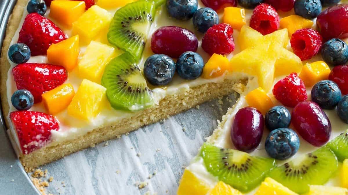 A round dessert with a cream-covered crust, topped with sliced strawberries, blueberries, kiwi slices, grapes, raspberries, and pieces of mango. One wedge-shaped slice has been removed.