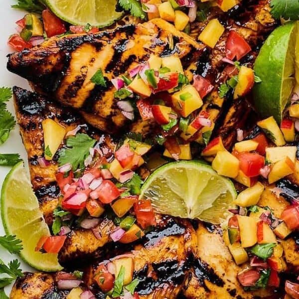 Grilled chicken breasts topped with a peach salsa, garnished with cilantro and lime wedges on a white plate.