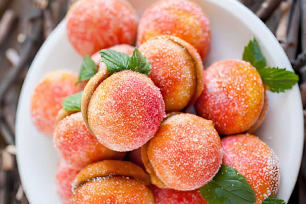 On a white plate are peach-shaped cookies covered with powdered sugar and adorned with small green leaves.