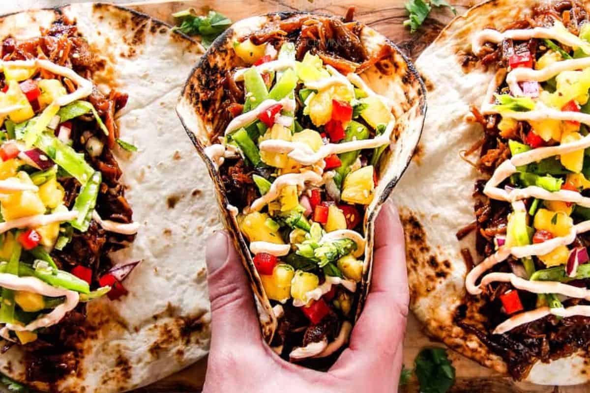 A hand holds a taco filled with shredded meat, diced pineapple, peppers, onions, lettuce, and drizzled sauce; two similar tacos rest on a wooden surface.