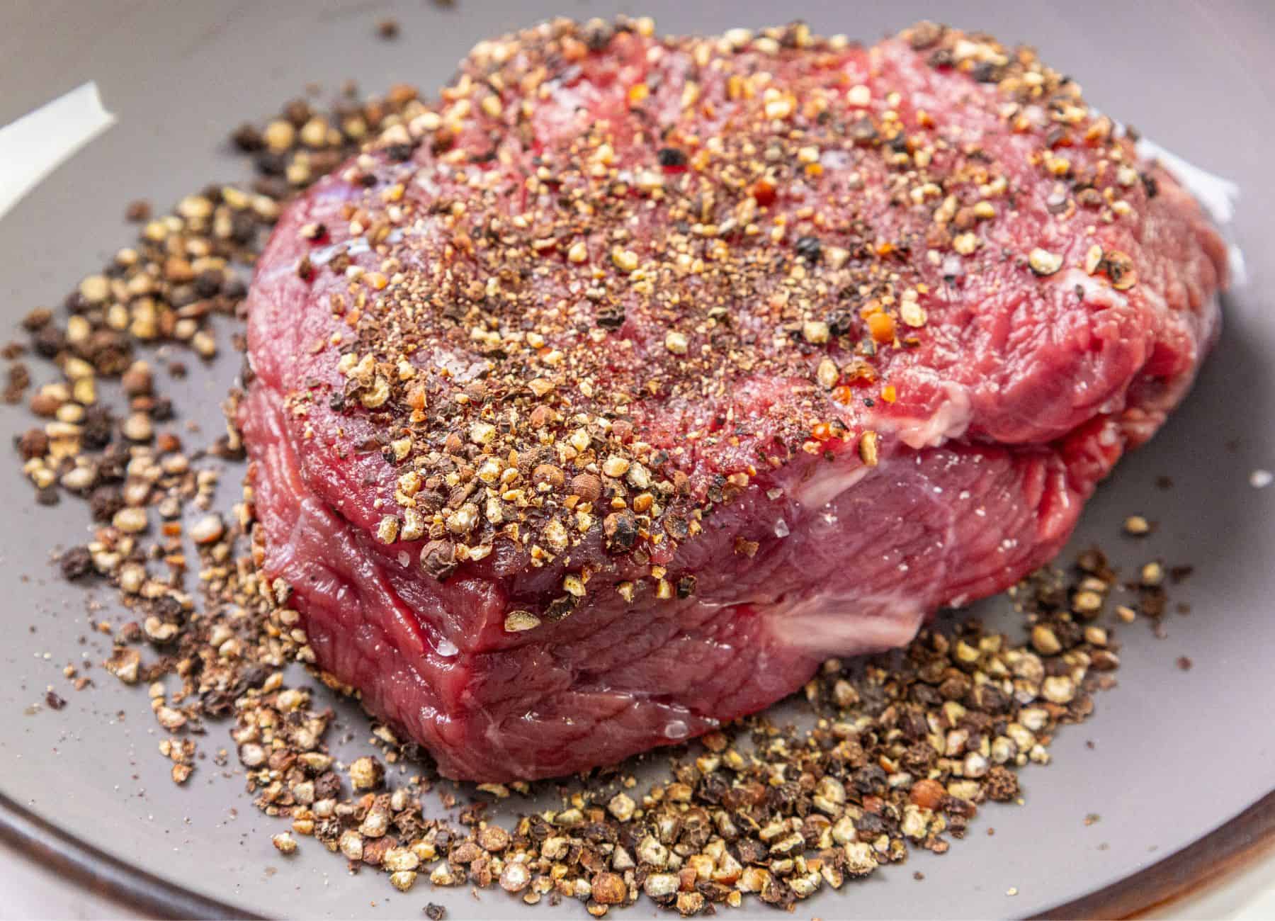 A raw steak coated with black pepper and spices on a gray plate, with loose pepper and seasoning scattered around it.