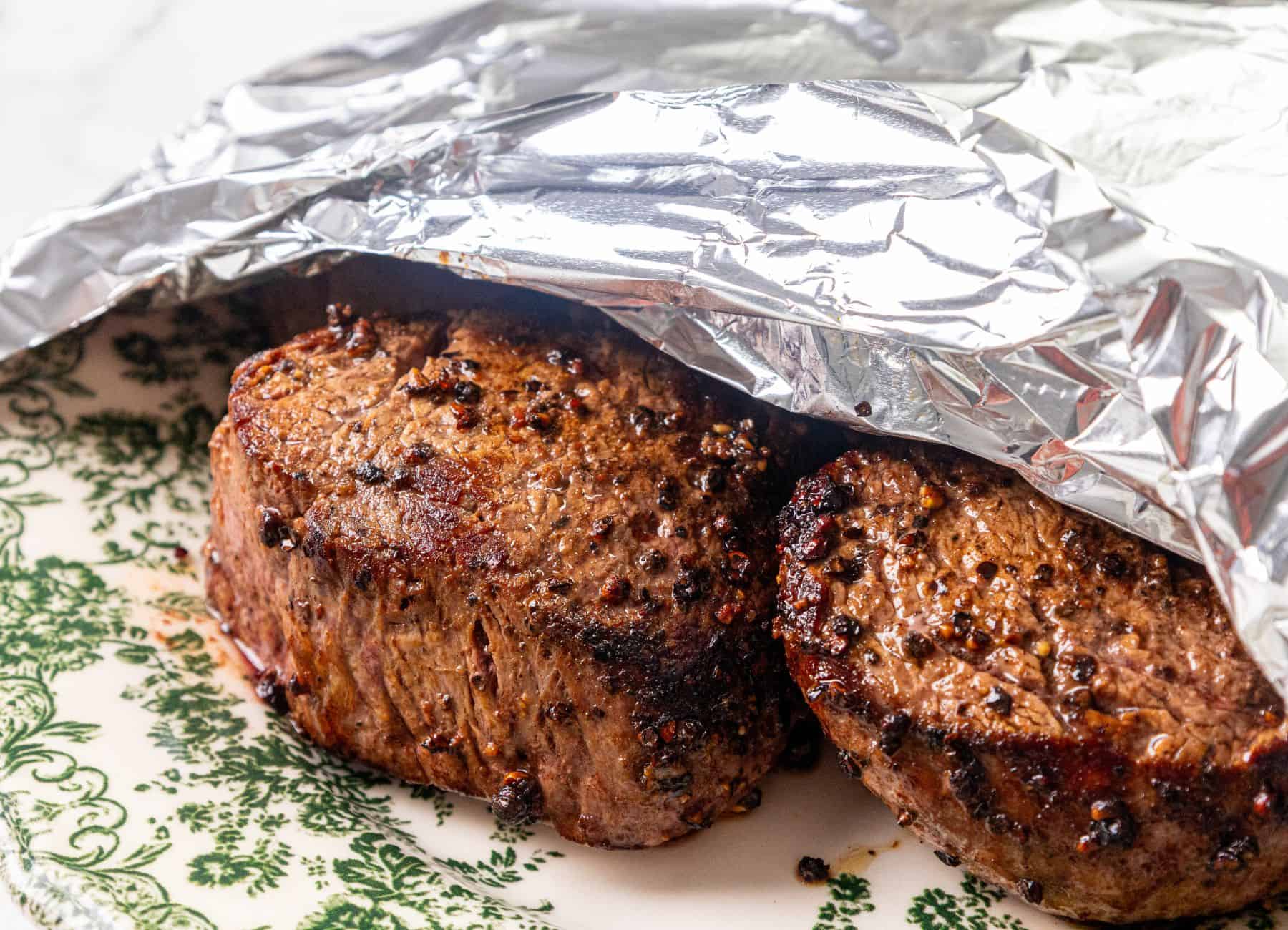 Two seared steaks on a decorative plate, partly covered by a piece of aluminum foil.