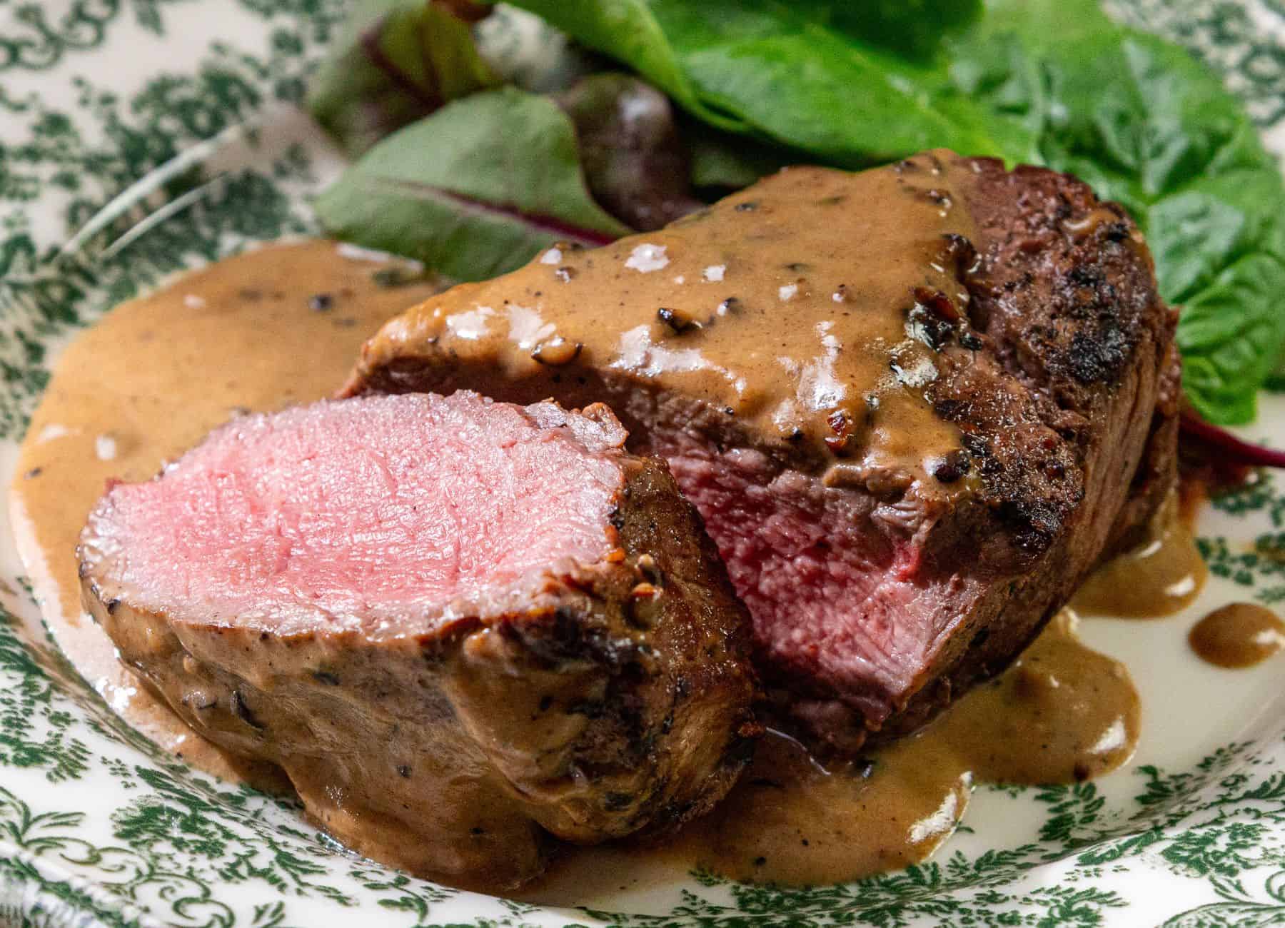 Sliced medium-rare steak with peppercorn sauce on a patterned plate, leafy greens in the background.