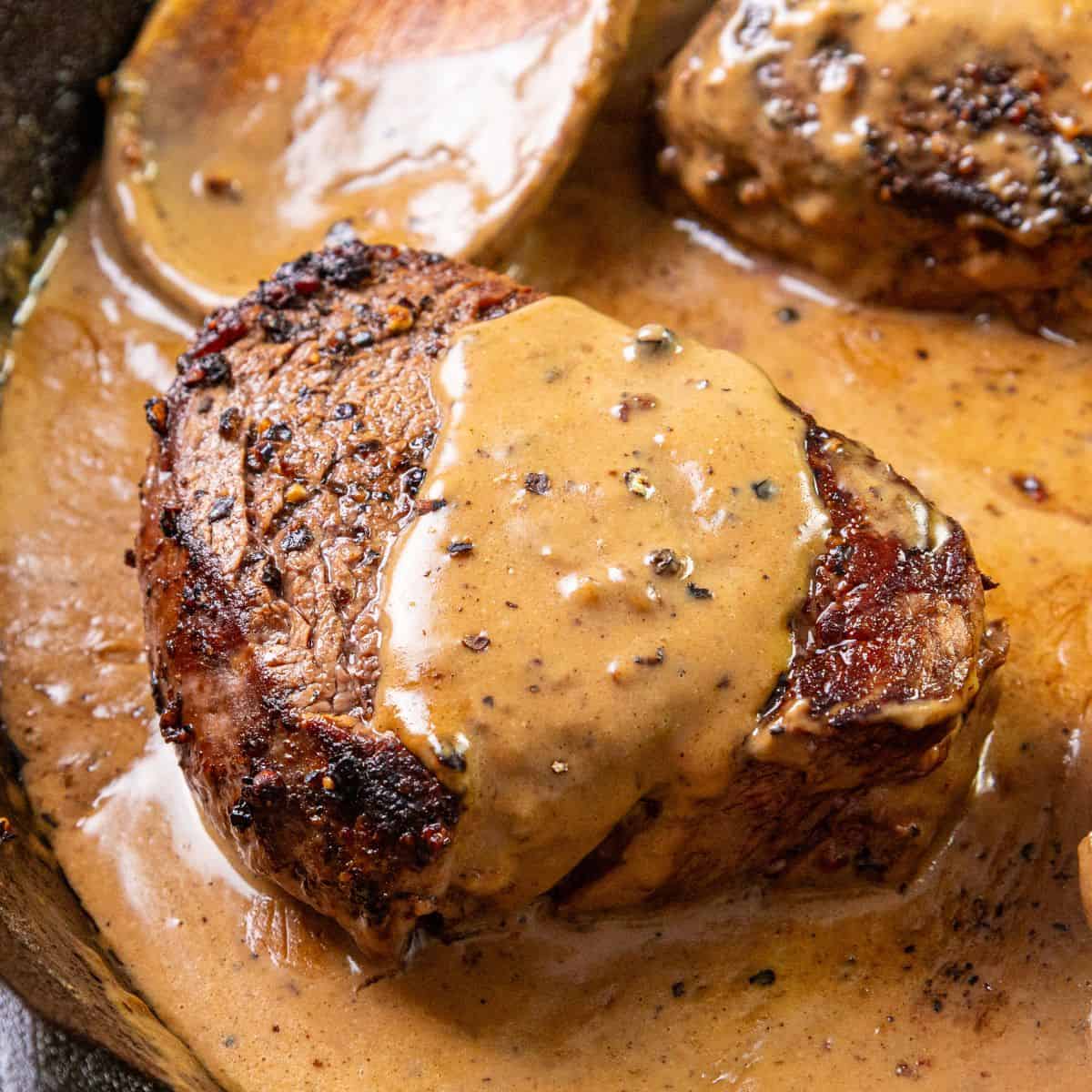 A seared steak topped with creamy peppercorn sauce sits in a skillet, with extra sauce displayed in the background.