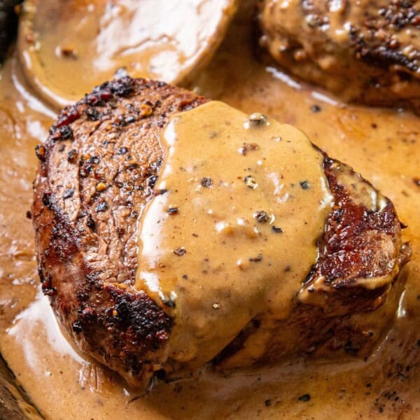 A seared steak topped with creamy peppercorn sauce in a skillet, with additional steak pieces partially visible behind.