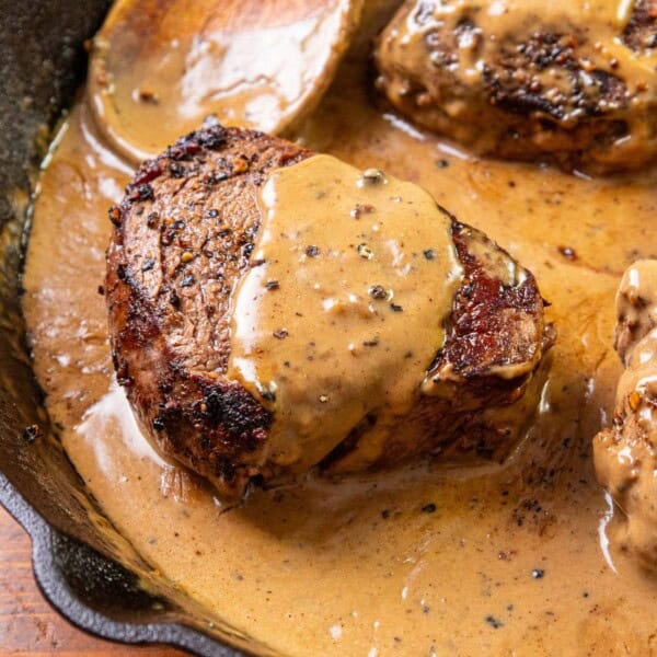 Three seared steaks in a cast iron skillet with creamy peppercorn sauce, black peppercorns, and fresh herbs on the side.