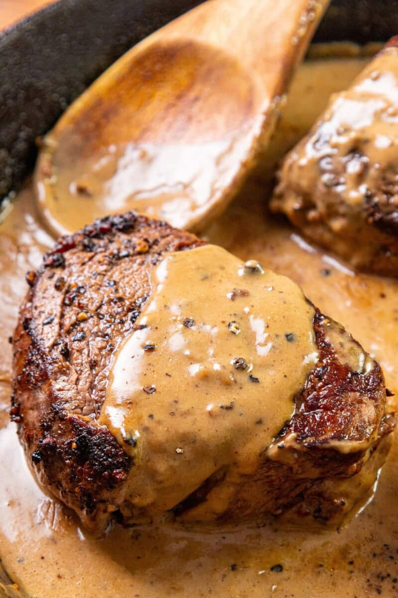 A cooked steak with creamy peppercorn sauce in a pan, wooden spoon resting to the side.