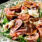 Fresh figs, prosciutto, mozzarella, arugula, and balsamic glaze arranged on a decorative plate with a fork beside it.