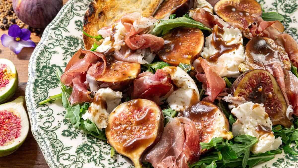Fresh figs, prosciutto slices, mozzarella, arugula leaves, and toasted bread with balsamic glaze on a patterned plate.