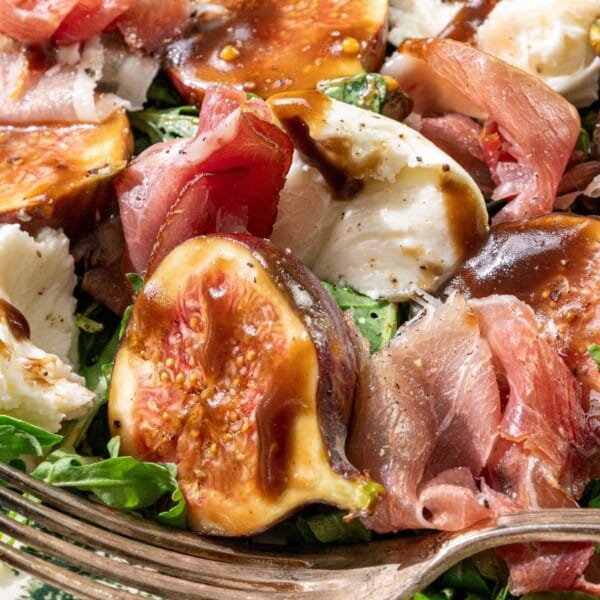 Fresh figs, prosciutto, mozzarella, arugula, and balsamic glaze arranged on a patterned plate with a fork.