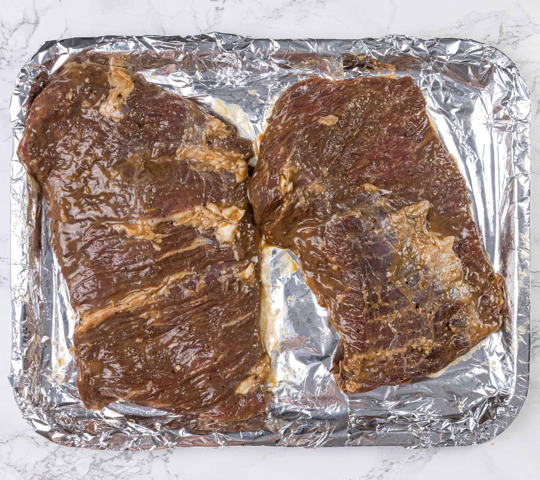 Two pieces of marinated raw steak sit on a foil-lined baking sheet.