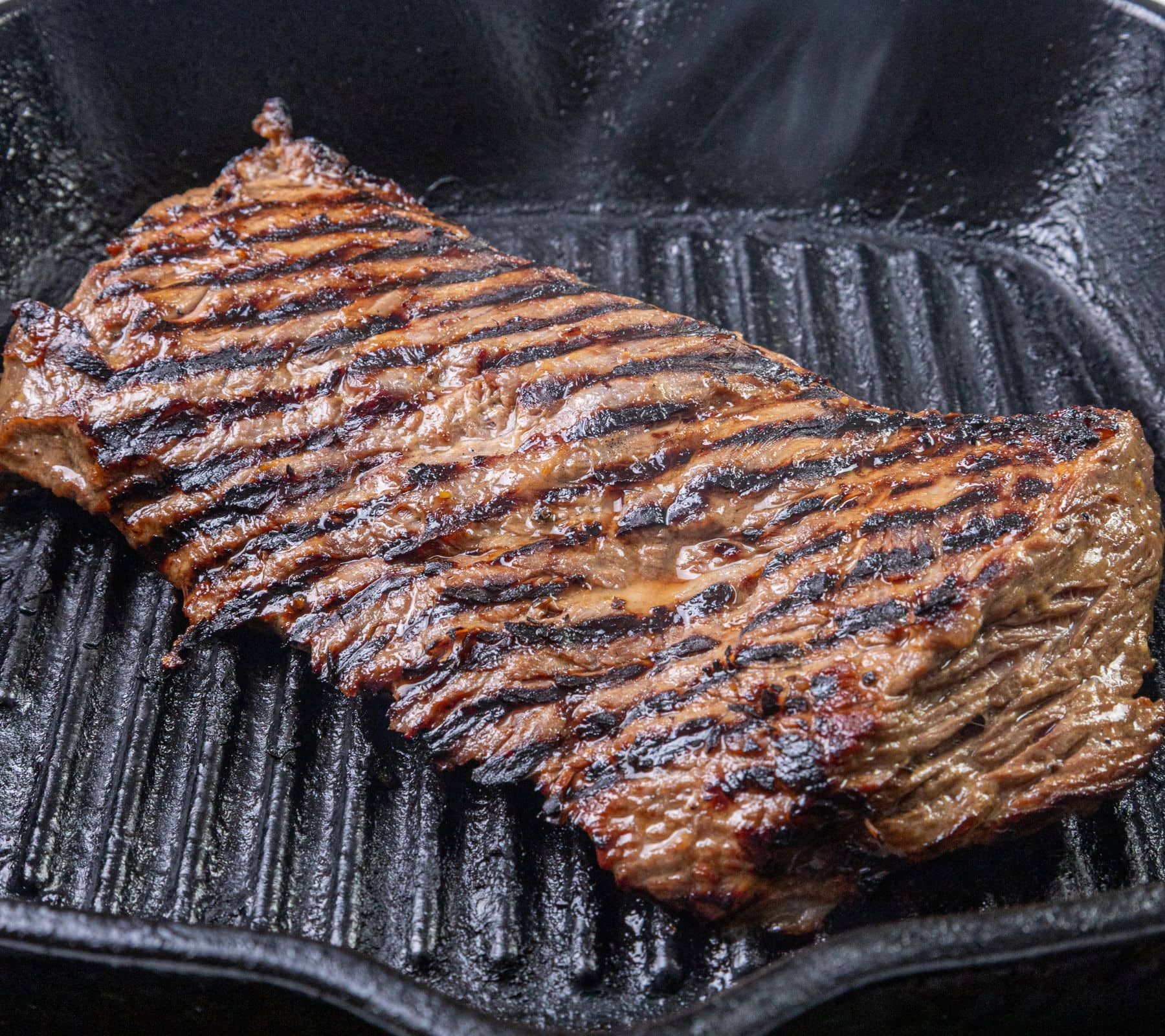 A grilled steak with char marks cooks in a black ridged cast iron grill pan.