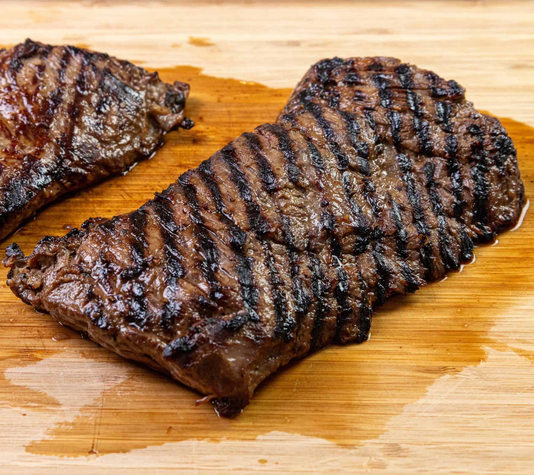 A grilled steak with dark sear marks sits on a wooden cutting board, with a smaller piece of steak partially visible next to it.